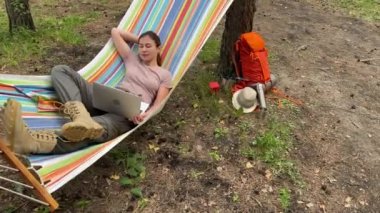 Ormandaki bir hamakta otururken dizüstü bilgisayarda çalışan beyaz bir kadın. Yürüyüşte kablosuz bilgisayar kullanan bir kız..