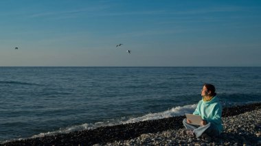 Bir çakıl taşı kumsalında otururken dizüstü bilgisayarda çalışan mutlu beyaz kadın..