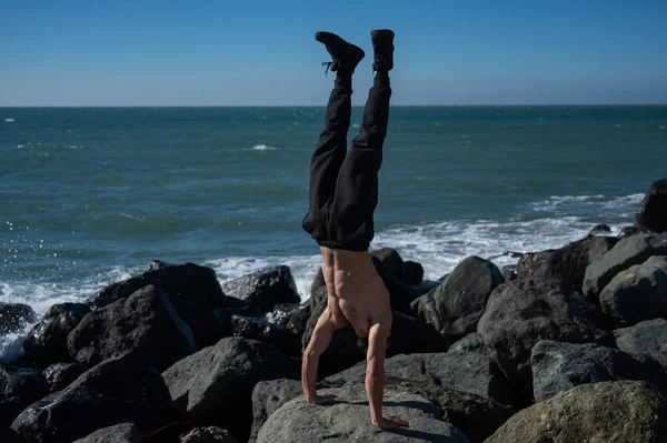 Uomo a torso nudo che fa stand su rocce in riva al mare. — Foto Stock