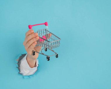 Avucunda alışveriş arabası olan bir kadın eli kağıt mavisi arka planda bir delikten dışarı sarkan.. 