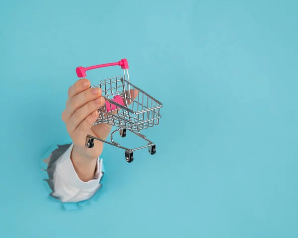 Mano de mujer con un carrito de compras en la palma que sobresale de un agujero en un fondo azul de papel. —  Fotos de Stock