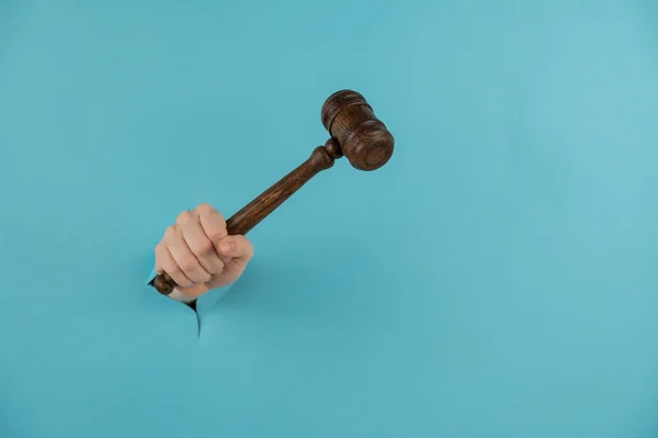 A womans hand with a wooden judges gavel sticks out of a hole in a blue background. Copy space. — Stock Photo, Image