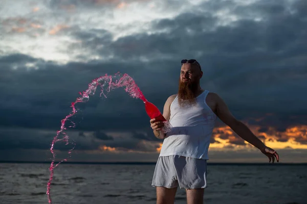 Юмористический портрет жестокого человека, наливающего газировку из бутылки на пляже на закате — стоковое фото