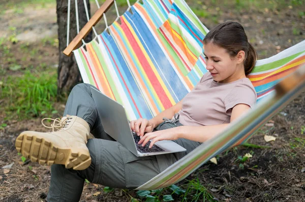 Mulher caucasiana trabalhando em laptop enquanto sentada em uma rede na floresta. Menina usa um computador sem fio em uma caminhada. — Fotografia de Stock