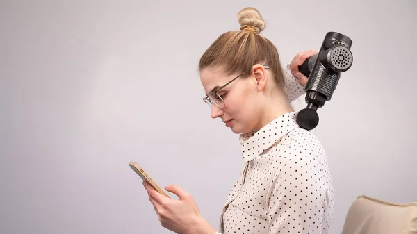 Una joven se sienta en una silla, usa un teléfono inteligente y se hace un masaje con una pistola de electro masaje. — Foto de Stock