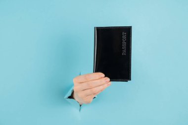 Bir kadının eli mavi bir kağıdın arkasındaki delikten dışarı çıkar ve siyah bir kapakta bir pasaport tutar.. 