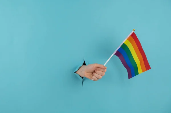 A womans hand sticks out of a hole in a blue paper background and holds an lgbt flag. — Stock Photo, Image
