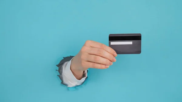 Een vrouwenhand die uit een gat in een papieren blauwe achtergrond steekt, heeft een creditcard.. — Stockfoto