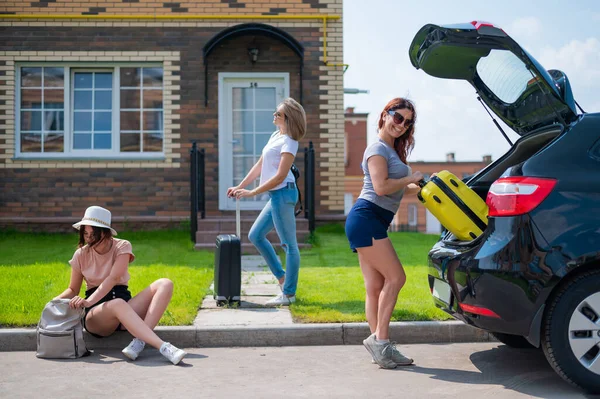 Drei kaukasische Frauen und ein Hund gehen auf Roadtrip. Die Mädchen laden ihre Koffer in den Kofferraum des Autos. Sommerferienkonzept mit Freundinnen. — Stockfoto