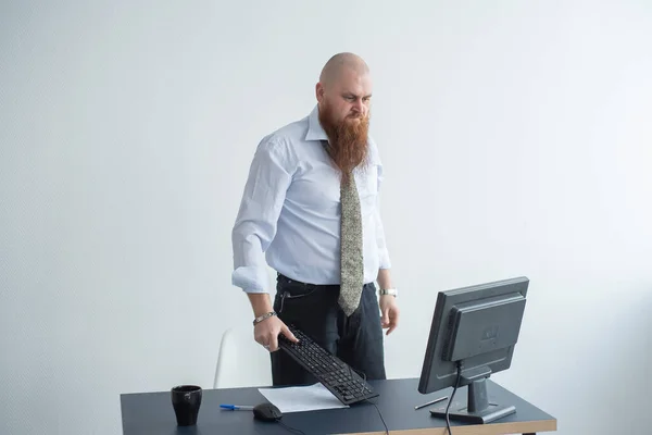 Un calvo enojado con barba roja en la oficina con traje de negocios choca contra una computadora. Manager con una crisis nerviosa rompe el teclado en la pantalla . —  Fotos de Stock