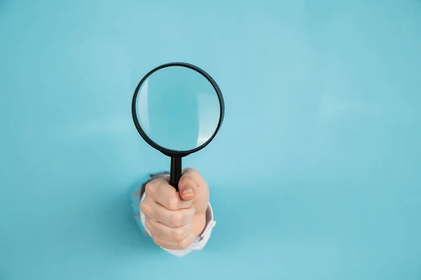 Una mano di donna che sporge da un buco in uno sfondo blu carta tiene una lente d'ingrandimento. Copia spazio. — Foto Stock