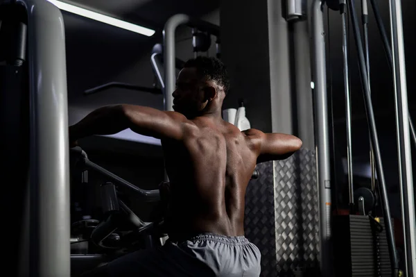 Póló nélküli afro-amerikai férfi hátgyakorlatokat végez egy gépen az edzőteremben.. — Stock Fotó