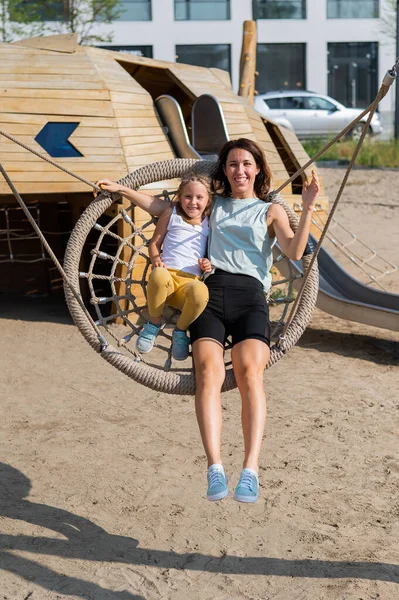 Mamma e figlia oscillano su un'altalena rotonda. Donna caucasica e bambina si divertono nel parco giochi. — Foto Stock