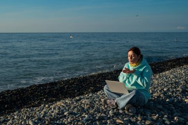 Beyaz kadın, çakıl taşlı plajda dizüstü bilgisayardan video ile iletişim kuruyor.. 