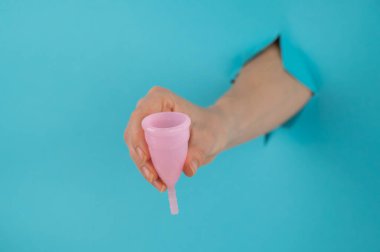 Mavi arka planda bir delikten dışarı sarkan bir kadın eli pembe bir adet bardağı tutuyor.. 