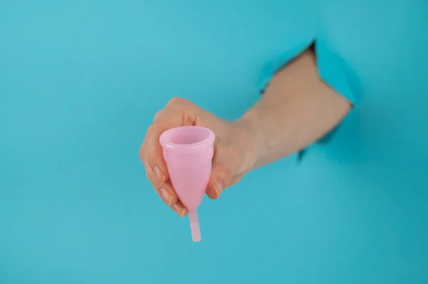 Egy kék hátterű lyukból kilógó női kéz rózsaszín menstruációs poharat tart.. — Stock Fotó