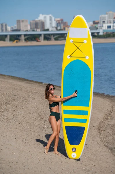 Mujer caucásica camina a lo largo de la playa y lleva un sup board en el río en la ciudad. Deportes de verano. —  Fotos de Stock