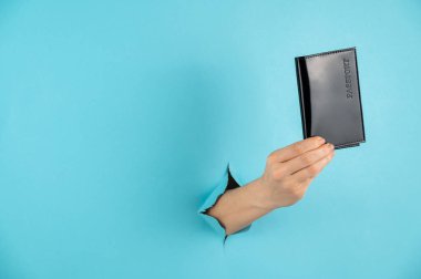 Bir kadının eli mavi bir kağıdın arkasındaki delikten dışarı çıkar ve siyah bir kapakta bir pasaport tutar.. 