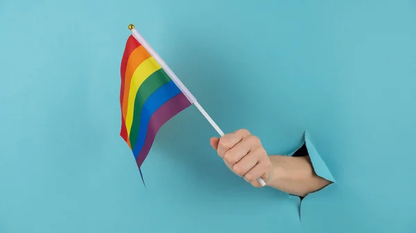 Una mano de mujer sobresale de un agujero en un fondo de papel azul y sostiene una bandera lgbt. — Foto de Stock