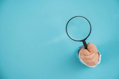 Kağıt mavisi arka planda bir delikten dışarı sarkan bir kadının eli büyüteç tutuyor. Boşluğu kopyala. 