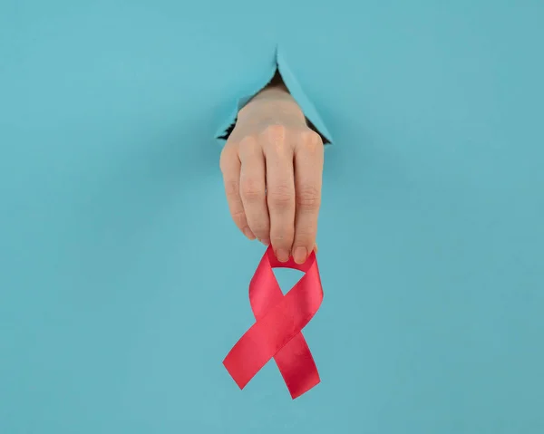 Kobieca ręka z czerwonym łukiem wystaje z dziury w niebieskim tle. Symbol raka piersi. — Zdjęcie stockowe