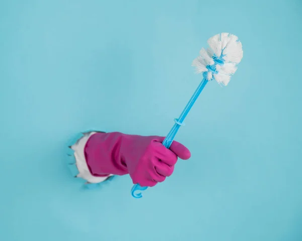 Una mano de mujer en un guante de goma sostiene un cepillo de inodoro y sale de un agujero sobre un fondo de papel azul. — Foto de Stock
