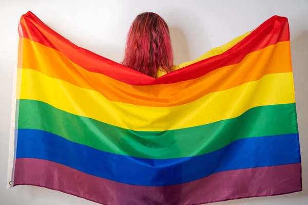 Young red-haired woman stands with her back against a white background and holds the lgbt flag — Stock Photo, Image