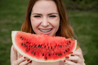 Diş telleri açık havada karpuz yiyen kızıl saçlı genç kadının yakın plan portresi.