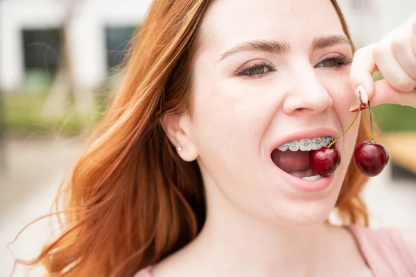 Hermosa joven pelirroja con tirantes en los dientes come cerezas dulces en el verano al aire libre —  Fotos de Stock