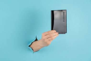 Bir kadının eli mavi bir kağıdın arkasındaki delikten dışarı çıkar ve siyah bir kapakta bir pasaport tutar.. 