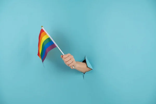 Una mano de mujer sobresale de un agujero en un fondo de papel azul y sostiene una bandera lgbt. —  Fotos de Stock