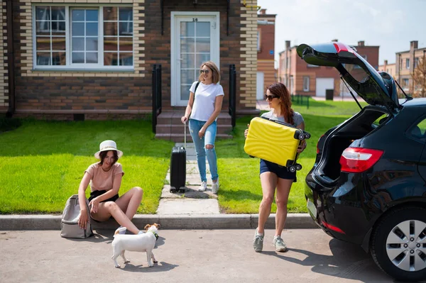 Drei kaukasische Frauen und ein Hund gehen auf Roadtrip. Die Mädchen laden ihre Koffer in den Kofferraum des Autos. Sommerferienkonzept mit Freundinnen. — Stockfoto
