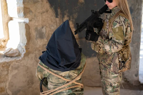 Una mujer caucásica con uniforme del ejército sostiene a un rehén con una bolsa en la cabeza al ver una ametralladora. — Foto de Stock