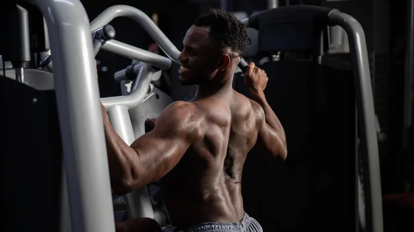 Shirtless african american man doing back exercises on a machine in the gym. — Stock Photo, Image