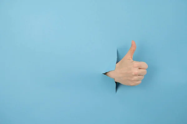 Egy kék hátterű lyukból kilógó női kéz hüvelykujjat mutat.. — Stock Fotó