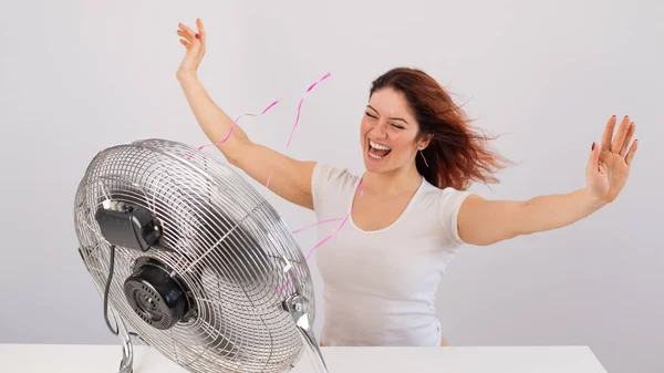 Joyful caucasian woman enjoying the wind blowing from an electric fan on a white background.
