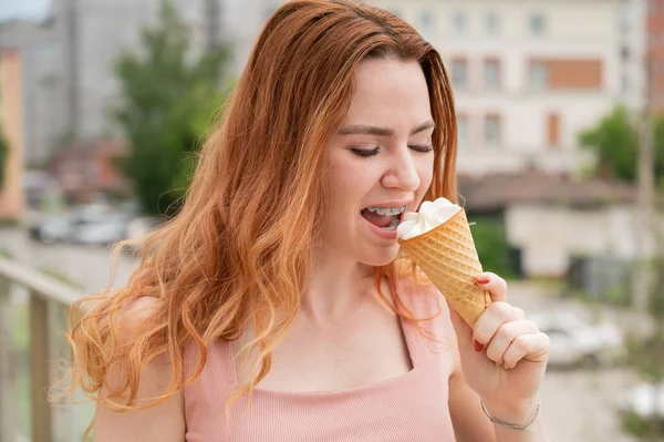 Ritratto di giovane bella donna dai capelli rossi che sorride con l'apparecchio e va a mangiare un cono gelato all'aperto in estate — Foto Stock