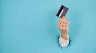 Bir kadının eli kağıtta bir delikten dışarı sarkan mavi arka planda bir kredi kartı tutar.