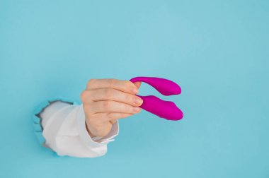 Bir kadının eli pembe bir seks oyuncağını kağıt mavisi bir arka planda bir delikten tutar.
