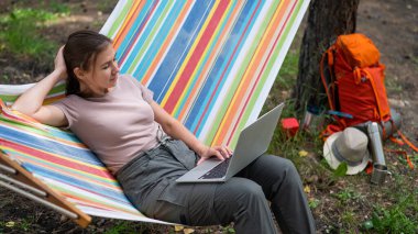 Ormandaki bir hamakta otururken dizüstü bilgisayarda çalışan beyaz bir kadın. Yürüyüşte kablosuz bilgisayar kullanan bir kız.