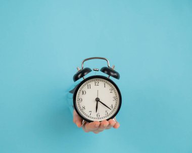 Bir kadın mavi bir kağıdın arkasındaki delikten dışarı sarkar ve çalar saati tutar.