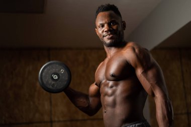 Çekici Afrikalı Amerikalı adam gülümsüyor ve halterlerle egzersiz yapıyor.