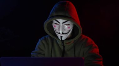 5 Haziran 2022 Novosibirsk, Rusya: Kapüşonlu Anonymous kırmızı ışıkta karanlıkta dizüstü bilgisayarda yazıyor