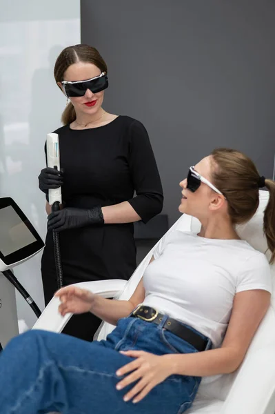 A woman on a couch in goggles undergoing a photorejuvenation procedure. Cosmetologist with a patient