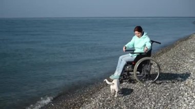 Tekerlekli sandalyedeki beyaz kadın deniz kenarında bir köpeğe sarılıyor.