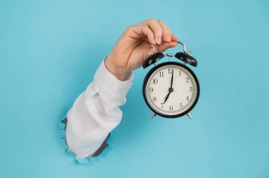 Bir kadın mavi bir kağıdın arkasındaki delikten dışarı sarkar ve çalar saati tutar.