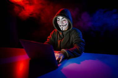 5 Haziran 2022 Novosibirsk, Rusya: Kapüşonlu Anonymous kırmızı-mavi dumanla karanlıkta dizüstü bilgisayarda yazıyor