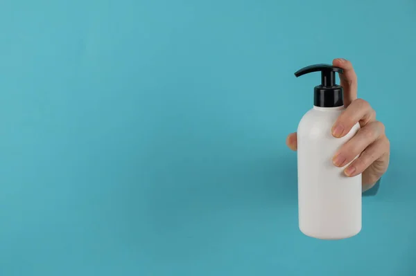 A womans hand sticks out of a hole in a blue paper background and holds a skin lotion pump