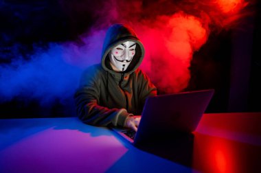 5 Haziran 2022 Novosibirsk, Rusya: Kapüşonlu Anonymous kırmızı-mavi dumanla karanlıkta dizüstü bilgisayarda yazıyor