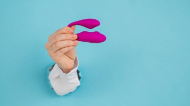 Bir kadının eli pembe bir seks oyuncağını kağıt mavisi bir arka planda bir delikten tutar.